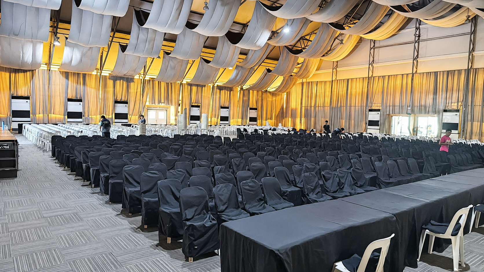 Black-covered chairs in theater rows beside black-skirted tables under gold and white ceiling draping