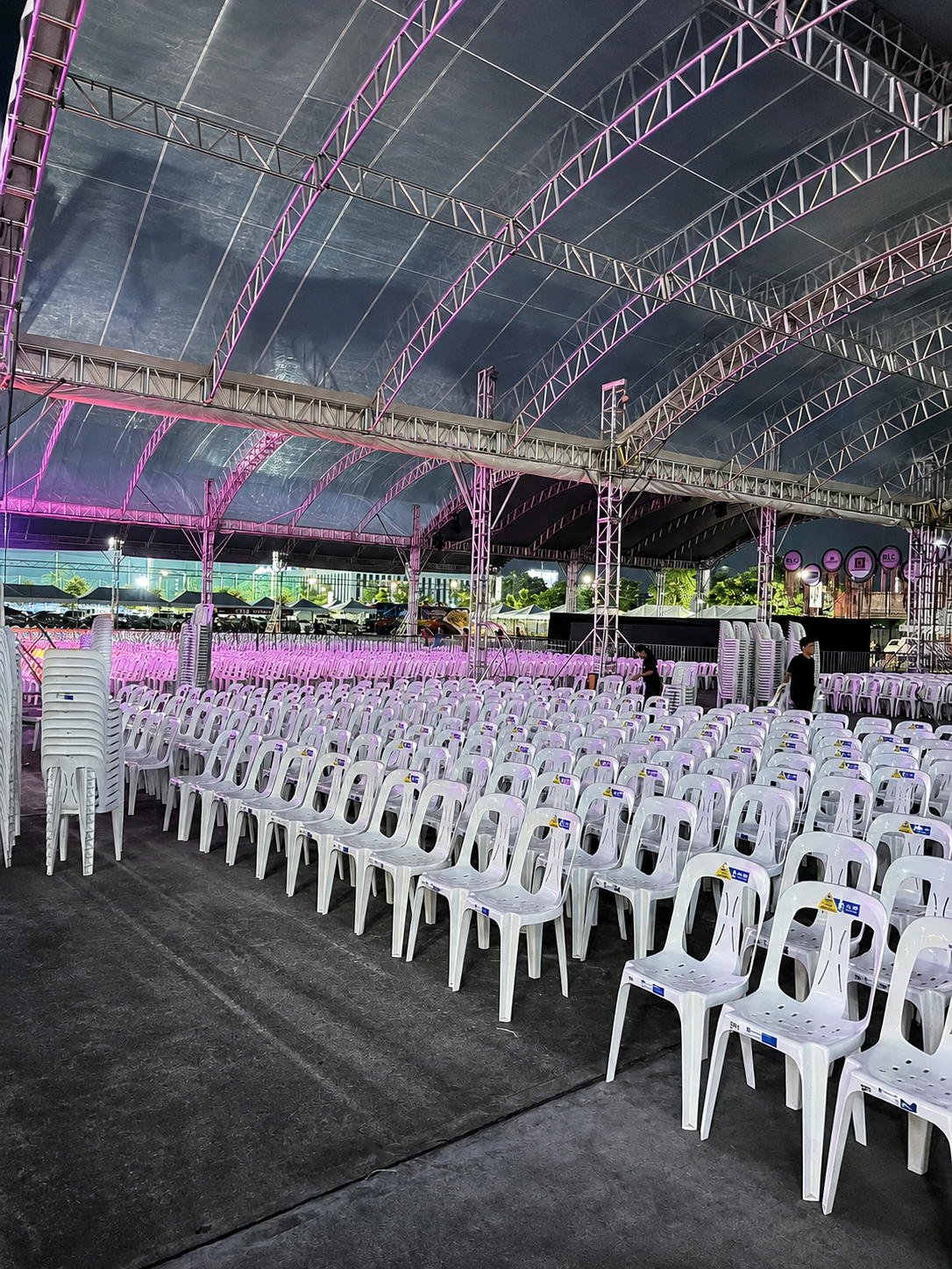 White monobloc chairs lined in rows under a trussed outdoor tent lit for an evening community program