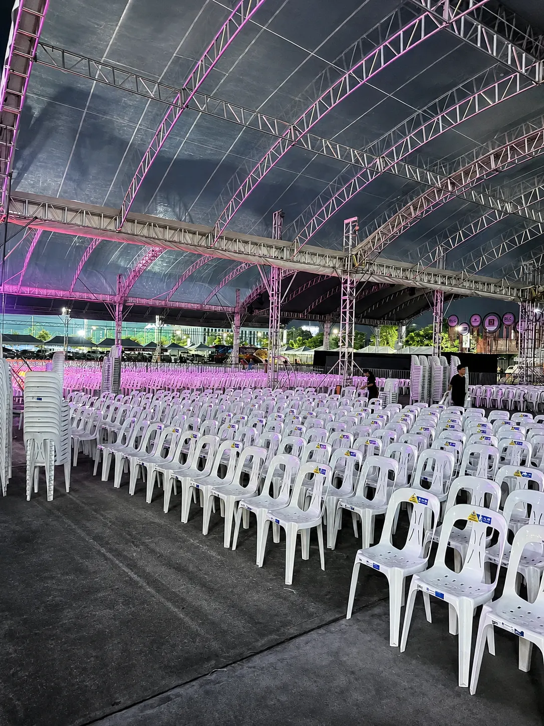 White monobloc chairs lined in rows under a trussed outdoor tent lit for an evening community program