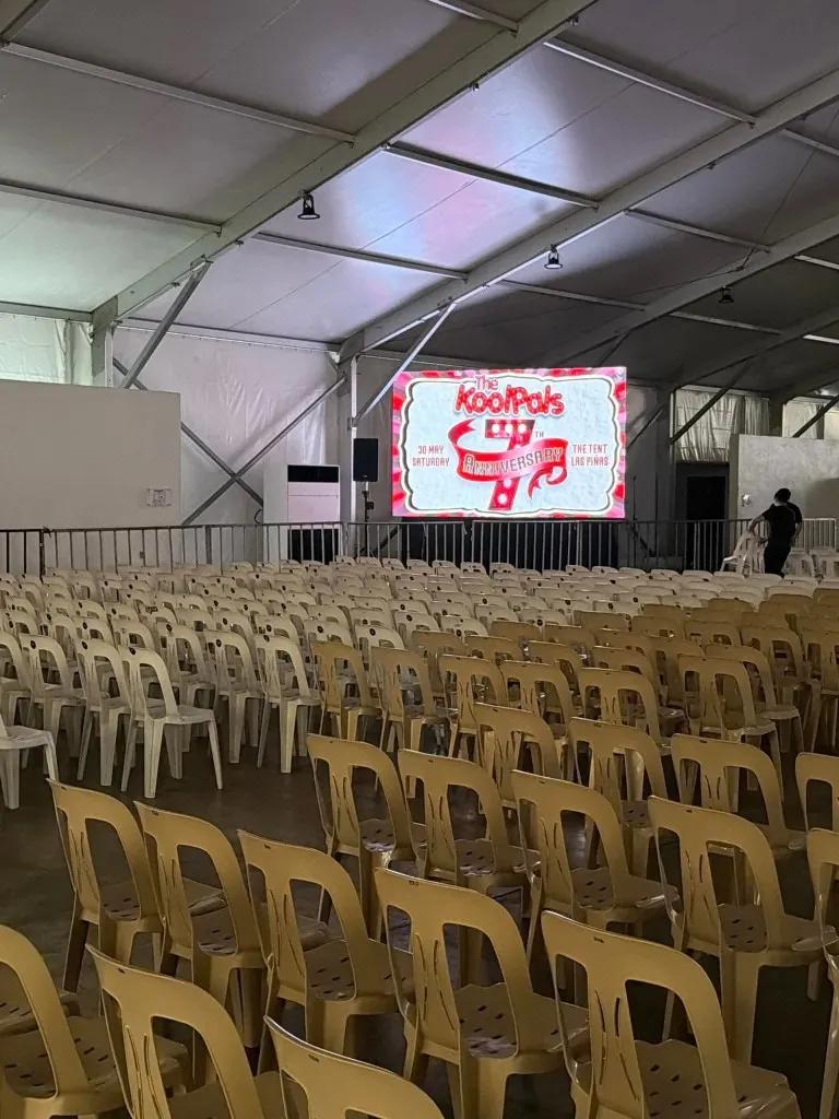 Rows of beige monobloc chairs inside a white event tent facing a large LED screen