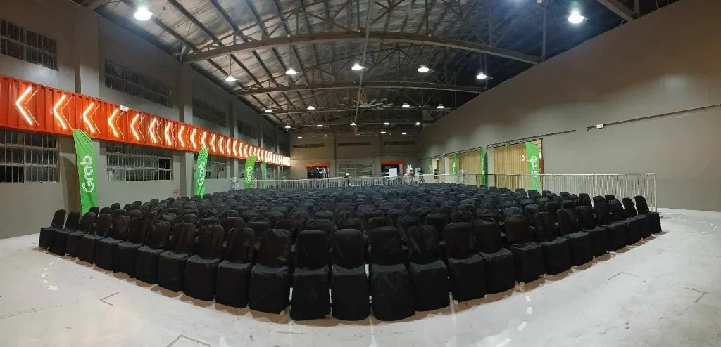 Hundreds of black-covered chairs set in theater rows inside a large industrial warehouse venue